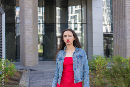 A young girl in a red dress stands in front of a building. She's wearing a denim jacket and red lipstick.