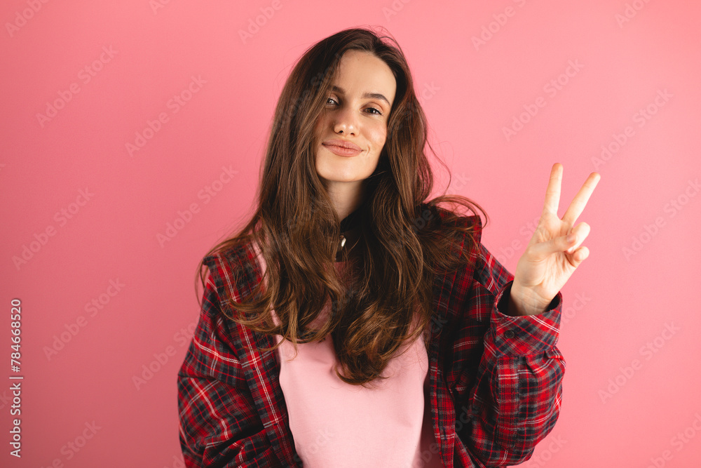 Smiling young woman gesturing peace sign against pink background. Brunette woman wear pink long sleeves top, plaid shirt. Happy laughing girl show v victory gestures, dancing, have fun and laughing. Smiling young woman gesturing peace sign against pink background. Brunette woman wear pink long sleeves top, plaid shirt. Happy laughing girl show v victory gestures, dancing, have fun and laughing.