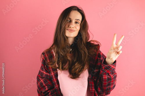 Smiling young woman gesturing peace sign against pink background. Brunette woman wear pink long sleeves top, plaid shirt. Happy laughing girl show v victory gestures, dancing, have fun and laughing.