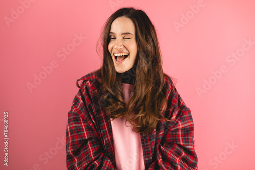 Playful happy contented brunette woman wear plaid shirt blinking eye, looking at camera with toothy smile, winking and flirting, expressing optimism. Indoor studio shot isolated on pink background.