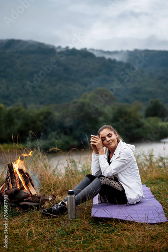 Travel, tourism, camping. Young calm woman tourist at the beautiful nature landscape sitting near the tent by the fire. Girl with drink in mug in her hands