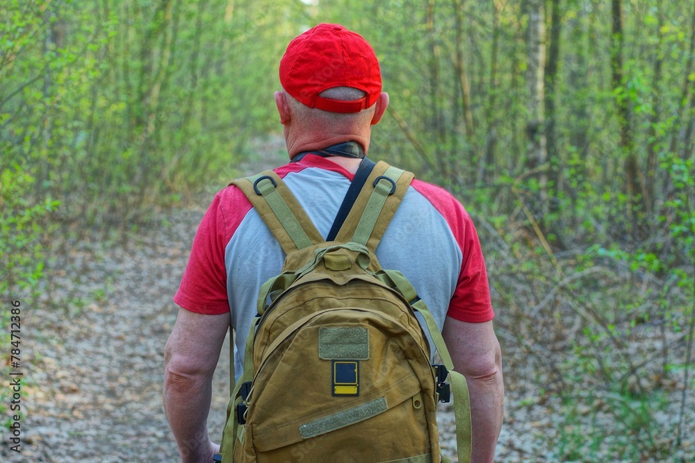 one male tourist in a red baseball cap with an olive-colored backpack on his back and dressed in a gray T-shirt walks along a road in the forest during the day one male tourist in a red baseball cap with an olive-colored backpack on his back and dressed in a gray T-shirt walks along a road in the forest during the day