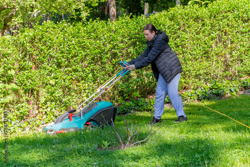 A middle-aged woman cuts the grass on her property with an electric lawnmower on a sunny day.