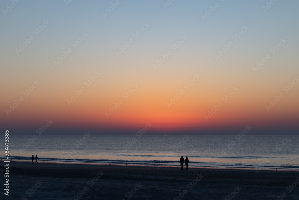 Figures in silhouette watch the sun rise over the ocean, very early morning beach walkers, horizontal aspect