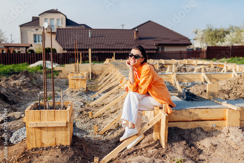 Happy woman owner sitting on concrete foundation for new house surrounded with wooden decking waiting for future home