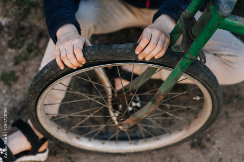 A teenage girl, a child, sits near an old retro bicycle with a broken, punctured wheel outdoors, pressing her hands on the tire. Close-up photography, portrait, lifestyle.