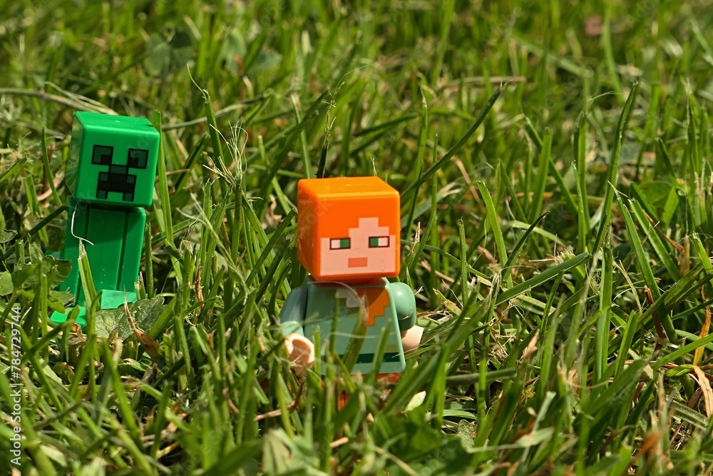 Naklejka premium LEGO Minecraft figure of female hero Alex escaping across freshly mowned garden lawn from green explosive Creeper mob. Spring daylight sunshine. 