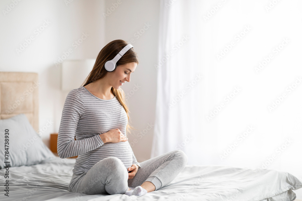 Pregnant lady seated on bed caressing her belly - obrazy, fototapety, plakaty 