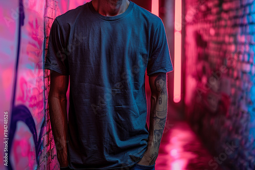 Mock up black t-shirt for logo, text or design. Young man with tattoo wears t-shirt. He stands on a brick wall background with with graffiti and neon lights