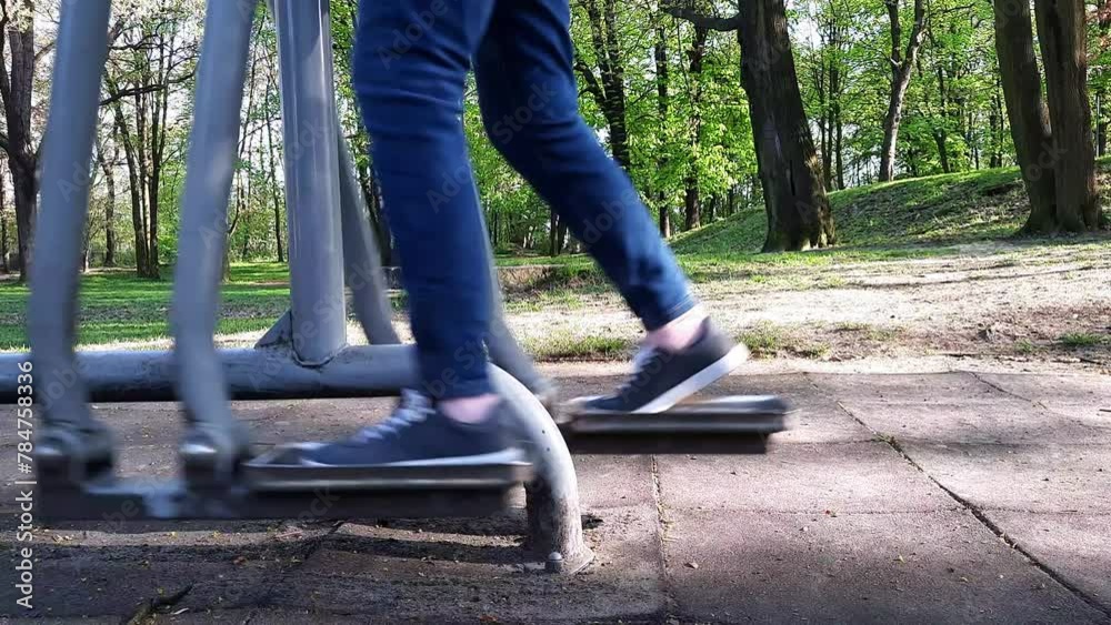 Person is training on Outdoor exercise machine air walker. Senior woman engages in outdoor fitness at park. Legs in jeans and moccasins close-up. Exercise and outdoor activities for pensioners.