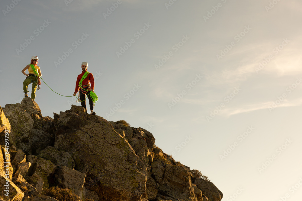 Fototapeta premium Two people are standing on a mountain, one of them wearing a red shirt. The other person is wearing a green harness. The scene is peaceful and serene, with the sun shining brightly in the background