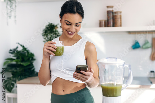 Pretty woman using her mobile phone while drinking fruit detox juice in the kitchen at home.