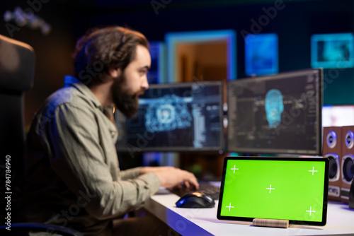 Focus on green screen device next to man in blurry background updating artificial intelligence neural networks on PC. IT admin upgrading high tech AI, mockup tablet on desk, close up
