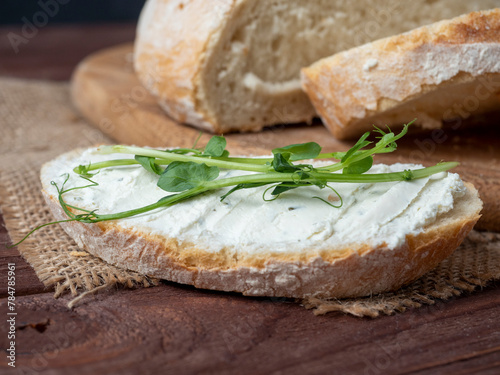 Close-up of a piece of bread smeared with cottage cheese and decorated with micro greenery. Next to fresh bread, linen napkin, wooden brown background. Side view
