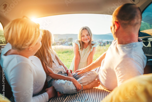  Portrait of happy smiling little girl gazing at camera. Happy young couple with two daughters inside the car trunk during auto trop. They laughing and chatting. Family values, traveling concept