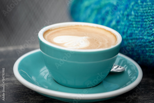 A close up ,blue, steaming cup of latte with artful foam, a heart, in a teal cup, accompanied by a cozy blue knit background.