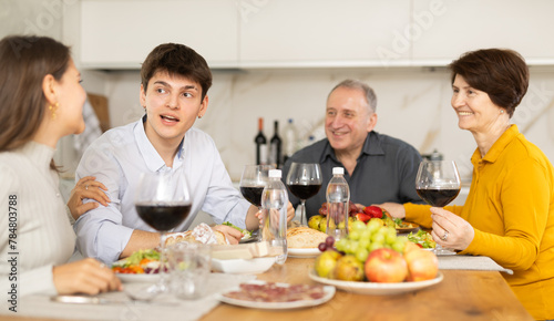 Smiling young guy engaging in lively conversation with sister and elderly parents at family table with wine and fruits in cozy home kitchen..