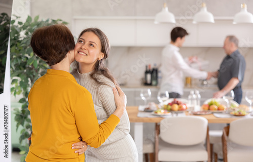 Happy young adult daughter warmly welcoming with hug and kiss senior mother coming to cozy family get-together
