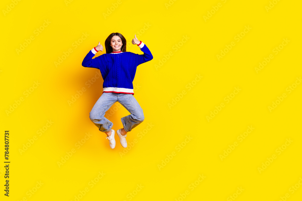 Full length photo of pretty teen girl jumping thumb up wear trendy knitwear blue outfit isolated on yellow color background