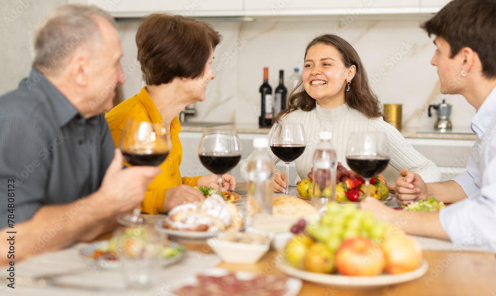 Positive daughter with boy-friend or husband and parents friendly conversation at dinner table with wine and light snacks in cozy kitchen