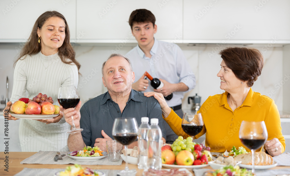 Happy carefree senior parents sharing memories with grown daughter and son-in-law during conversation over family celebration with wine at cozy home environment