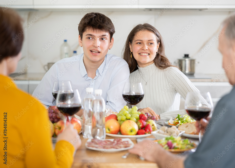 Smiling guy with wife visiting parents. Young couple sitting with glasses of wine at family holiday table