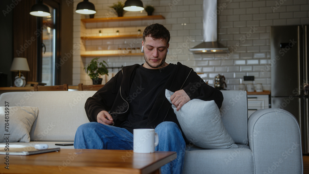 Young man sitting on the sofa and listening to music with wireless in-ear headphones and singing along to the music he is listening to. Man having fun, spending time alone at home