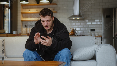  Young man sitting on sofa reacts to what sees, surfing internet on mobile phone, checking email, reading media news, scrolling social medias, using mobile applications on smartphone at home