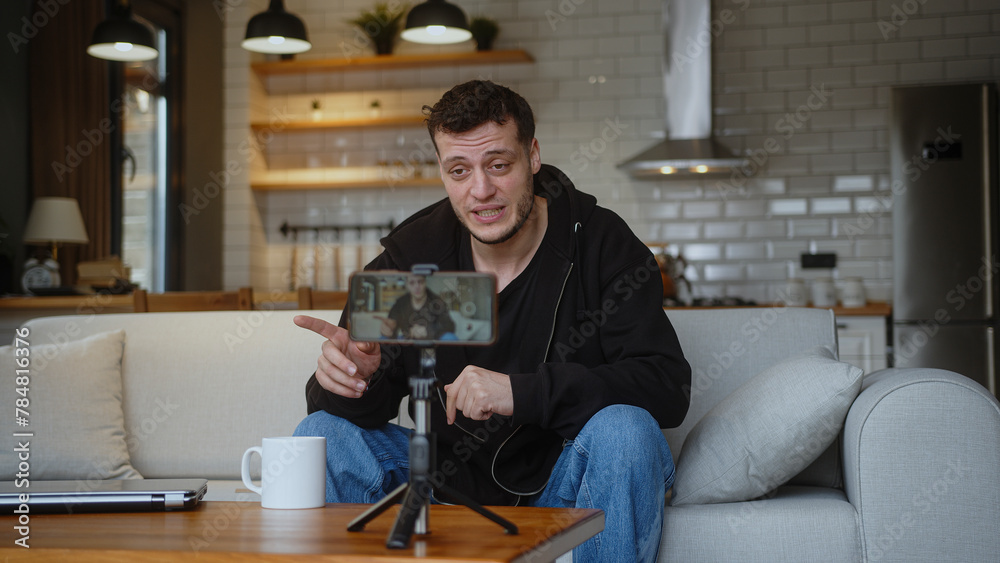 Influencer vlogger man sit on the couch recording video blog on mobile camera tripod talk for personal media channel, streaming live session blogging broadcast at home.