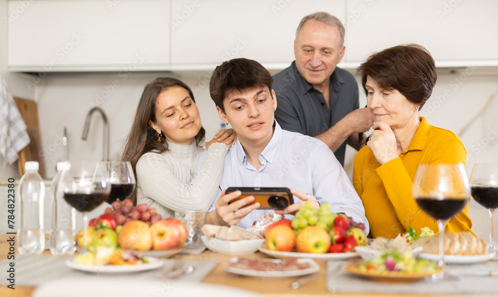 Positive young couple sharing memories with elderly parents sitting at cozy kitchen, browsing photos on smartphone together during festive family meal