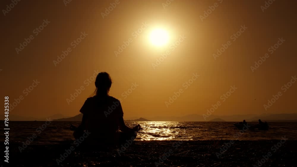 Girl doing calm stretch on dusk shore. A sporty girl doing calm yoga in lotus pose on the sea shore against bright evening in warm summer. Girl doing calm stretch on dusk shore. A sporty girl doing calm yoga in lotus pose on the sea shore against bright evening in warm summer.