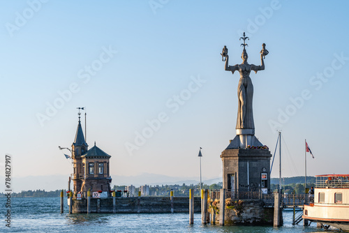 Die Imperia-Statue wacht neben dem Leuchtturm an der Hafeneinfahrt mit der Konstanzer Fahne im Wind in der Morgensonne. Konstanz, Bodensee, Baden-Württemberg, Deutschland, Europa.