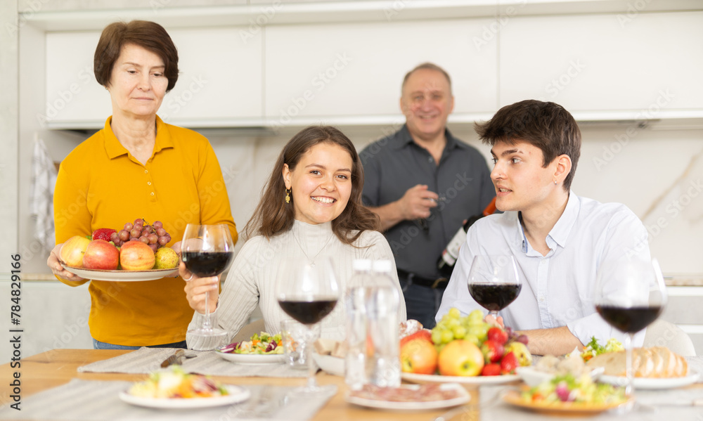 Smiling young guy with girlfriend visiting parents. Young couple sitting with glasses of wine at family holiday table
