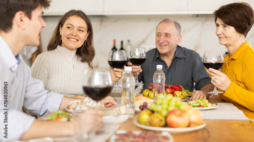 Positive daughter with boy-friend or husband and parents friendly conversation at dinner table with wine and light snacks in cozy kitchen