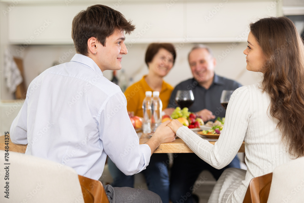 Young couple exchanging loving looks and holding hands while sitting at cozy set home table with smiling elderly parents watching approvingly, enjoying warm family atmosphere..