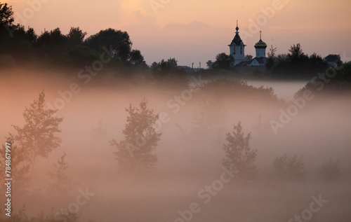 The church on the edge of the village in the evening pink fog during sunset