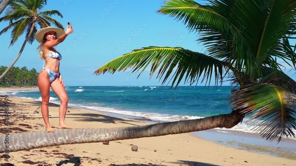 A sexy influencer in a bikini and a large white sun hat poses on the trunk of a coconut tree on a beautiful tropical beach and takes selfies with her smartphone. A sexy influencer in a bikini and a large white sun hat poses on the trunk of a coconut tree on a beautiful tropical beach and takes selfies with her smartphone.