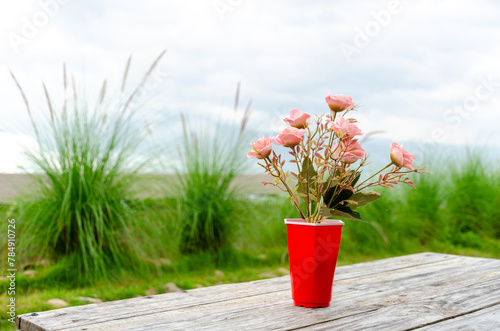 Pink rose flower in red vase on wooden table with nature background
