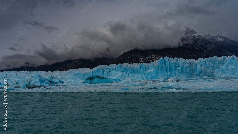 A wall of blue ice with cracks and sharp peaks stretches in a glacial lake. Thawed ice floes, icebergs float in turquoise water. Mountains in clouds and fog. Perito Moreno glacier. Lago Argentino.