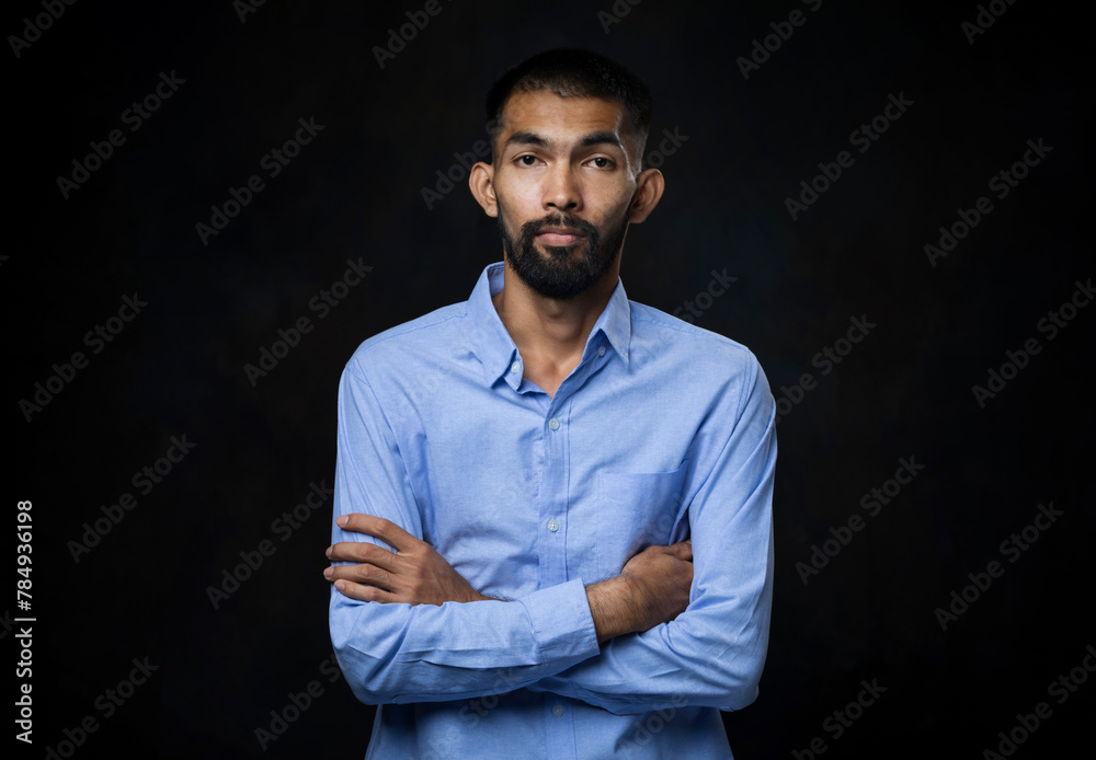 Fototapeta premium Portrait of young Asian man with beard in a blue shirt with his arms crossed and looking at camera in black studio background.