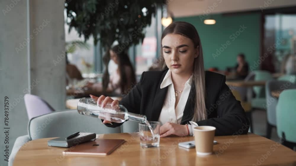 young, attractive woman sitting in a café, dressed in business attire, pours water from a bottle into a glass and drinks it young, attractive woman sitting in a café, dressed in business attire, pours water from a bottle into a glass and drinks it