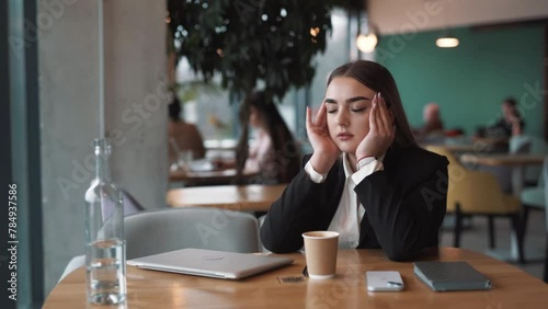 tired, beautiful woman sits in a stylish cafe in business attire, closing her laptop and finishing work with a headache, massaging her temples to relieve migraine pain photo