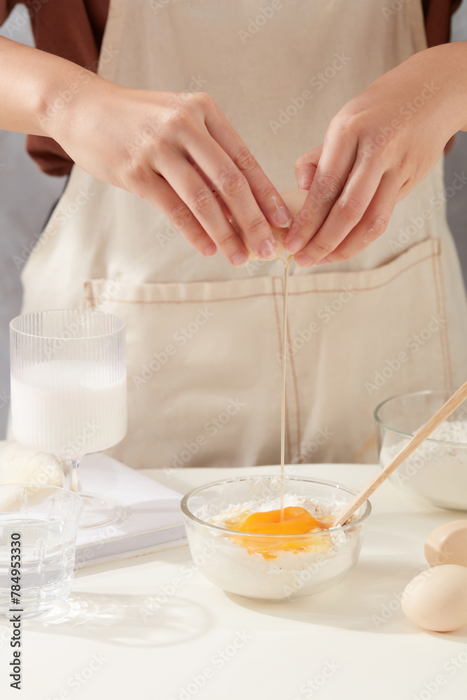 Baking ingredients concepts with female hands are cracking an egg in a bowl of flour to prepare homemade pastry on minimals white kitchen table next to ingredients. Front view photo Baking ingredients concepts with female hands are cracking an egg in a bowl of flour to prepare homemade pastry on minimals white kitchen table next to ingredients. Front view photo
