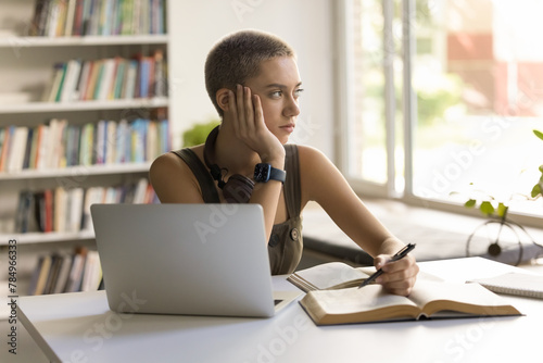 Εκτύπωση καμβά Thoughtful student girl sitting at desk with textbook and laptop, looks out wind