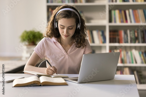 Attractive teenage student girl wear headphones studying in library, sit at desk with laptop, make assignment, writes essay, listen audio course, improve foreign language skills, studying in classroom