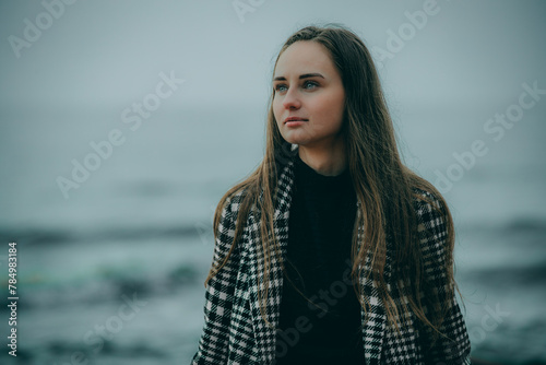 A woman is sitting on a rocky shore with waves crashing behind her. Black dress, checkered coat. Cloudy sky.