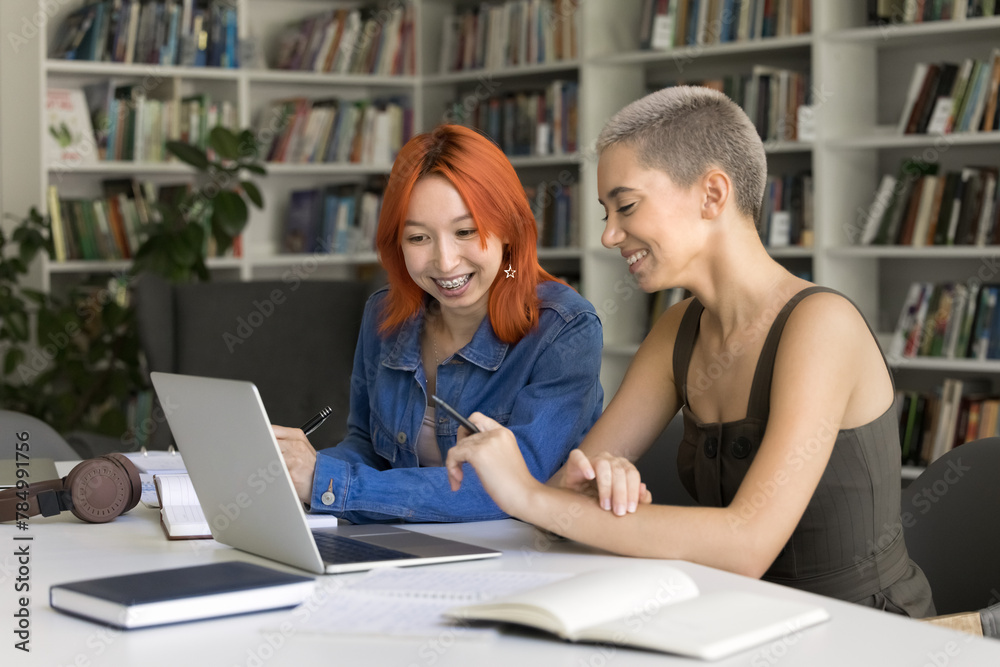 Two pretty redhead and blonde schoolmates working on joint on-line project, sit at desk, look at laptop, watching webinar, make research, engaged in successful friendly teamwork in high-school library Two pretty redhead and blonde schoolmates working on joint on-line project, sit at desk, look at laptop, watching webinar, make research, engaged in successful friendly teamwork in high-school library