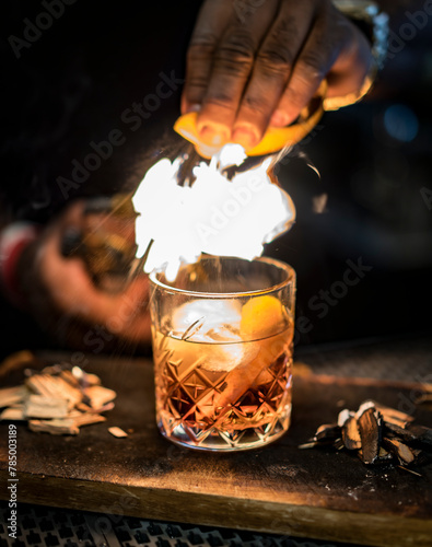 Old fashioned Whiskey drink made by Bartender with fire torched orange peel zest in a smoked wood rocks glass.