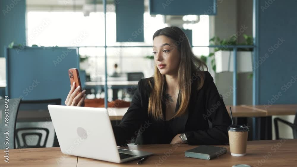 cheerful, beautiful young woman sits in a stylish coworking office in business attire, happily greeting someone via video call while talking on her smartphone with a laptop on the table cheerful, beautiful young woman sits in a stylish coworking office in business attire, happily greeting someone via video call while talking on her smartphone with a laptop on the table