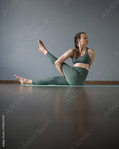 Young woman working out in gym on mat. ardha matsyendrasana. Asana sitting on floor.Holding straight leg while sitting on floor. Yoga trainer. Stretching. Gray background. Reflective floor. Side view. photo